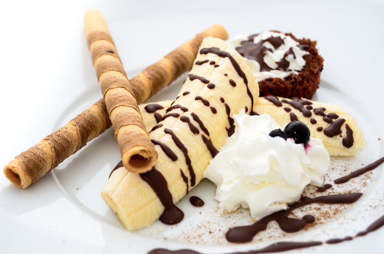 services-03 Close-up of a banana dessert with chocolate drizzle, whipped cream, and wafer sticks.